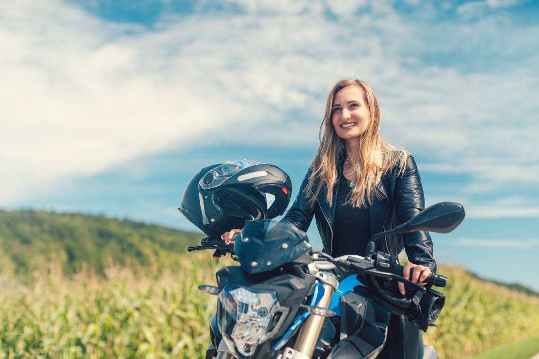 A woman in a leather jacket smiling while sitting on a motorcycle outdoors in front of a field beneath a blue sky.