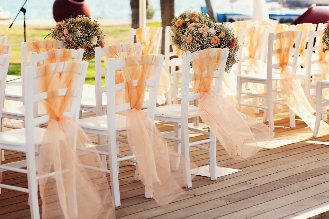 Multiple rows of white chairs with ribbons and bouquets tied to them. The chairs are on a wooden deck at an outdoor wedding.