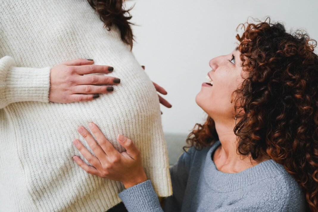 A woman holding her pregnant belly while another woman kneels down to hold her belly and look up at her excitedly.