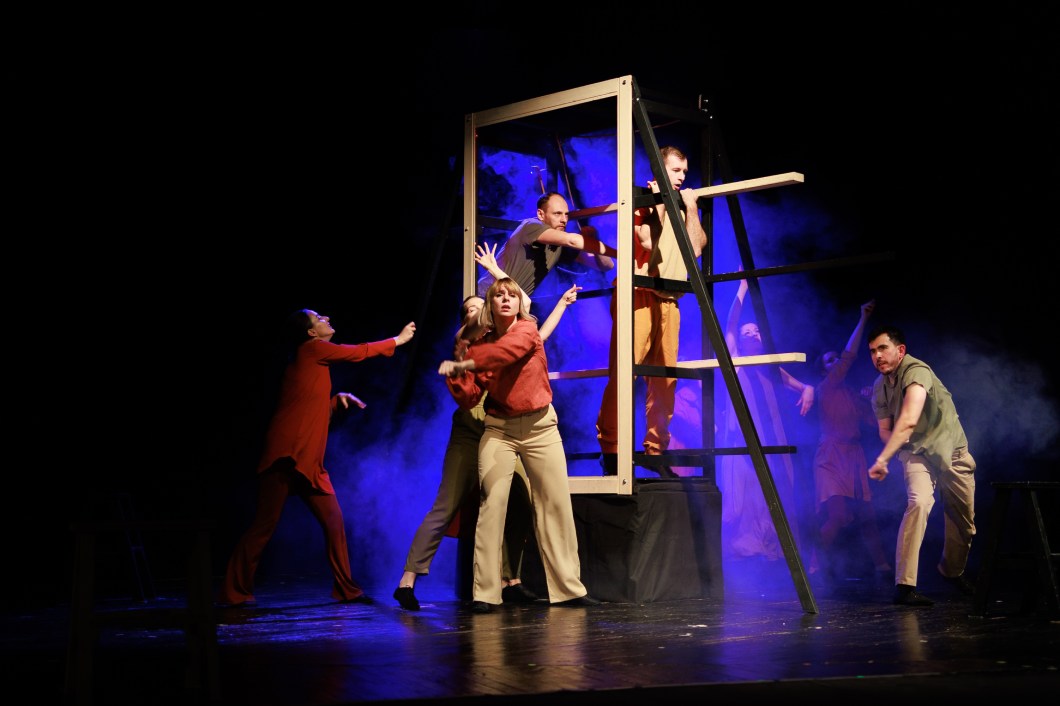 A cast of actors and actresses performs a play on a darkened stage. It features a minimal set that includes a frame.